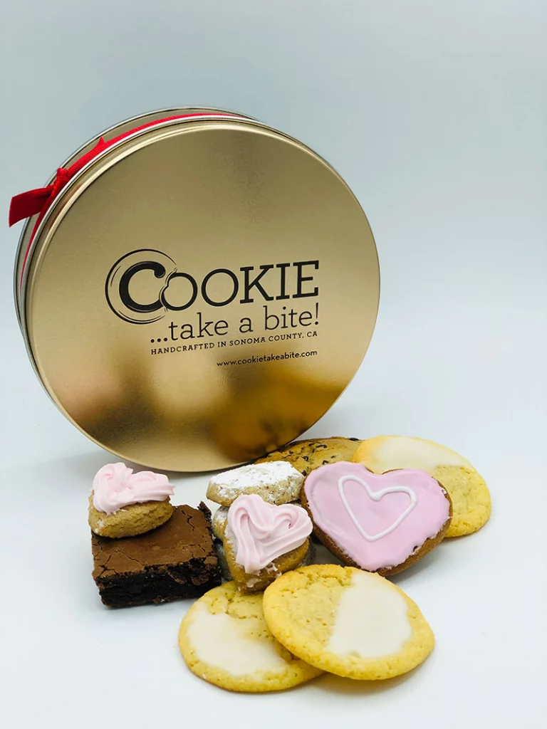 Cookie...take a bite! Premium Mother's Day Tin with a variety of cookies in front including pink heart-shaped cookies.