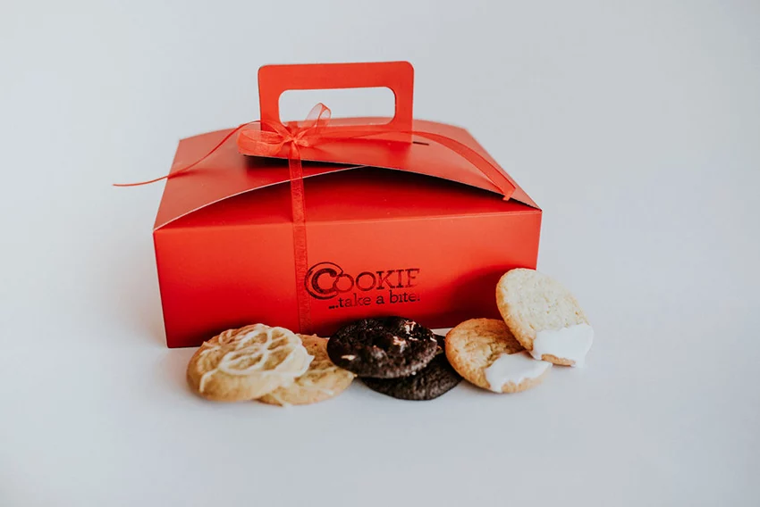 A red box with the COOKIE... take a bite! logo stamped on it with a variety of fresh baked cookies in front of it