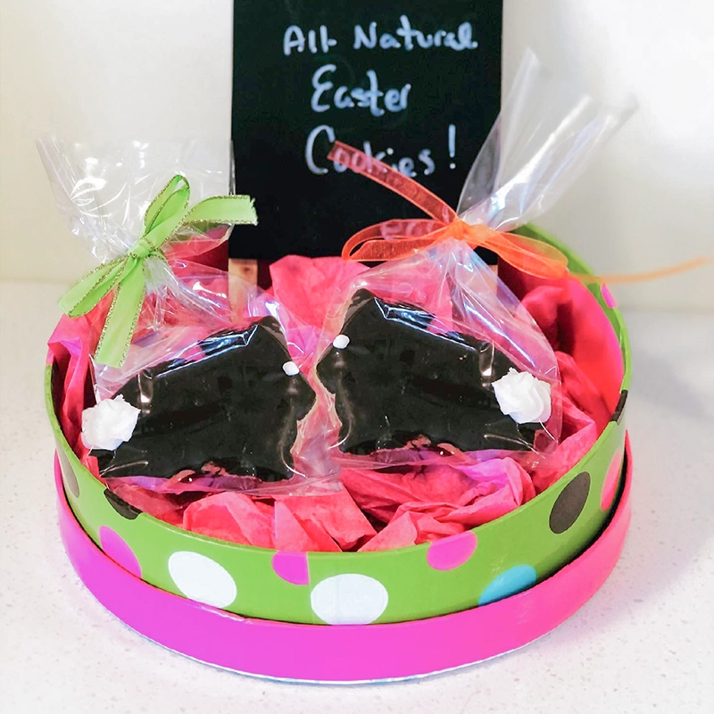 Chocolate covered graham cracker Easter bunny cookies in a gift tin with brightly colored tissue paper and a chalkboard sign in the background that reads, "All-Natural Easter Cookies!"