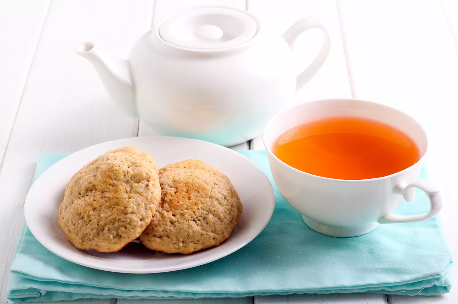 Cup of reddish tea in tea cup with saucer of two handmade cookies. Small tea pot behind.