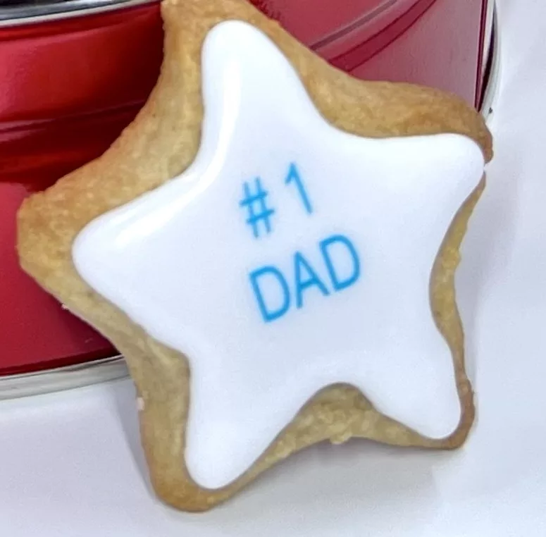 Star-shaped cookie with white icing and #1 DAD printed in blue standing up against a cookie tin. 