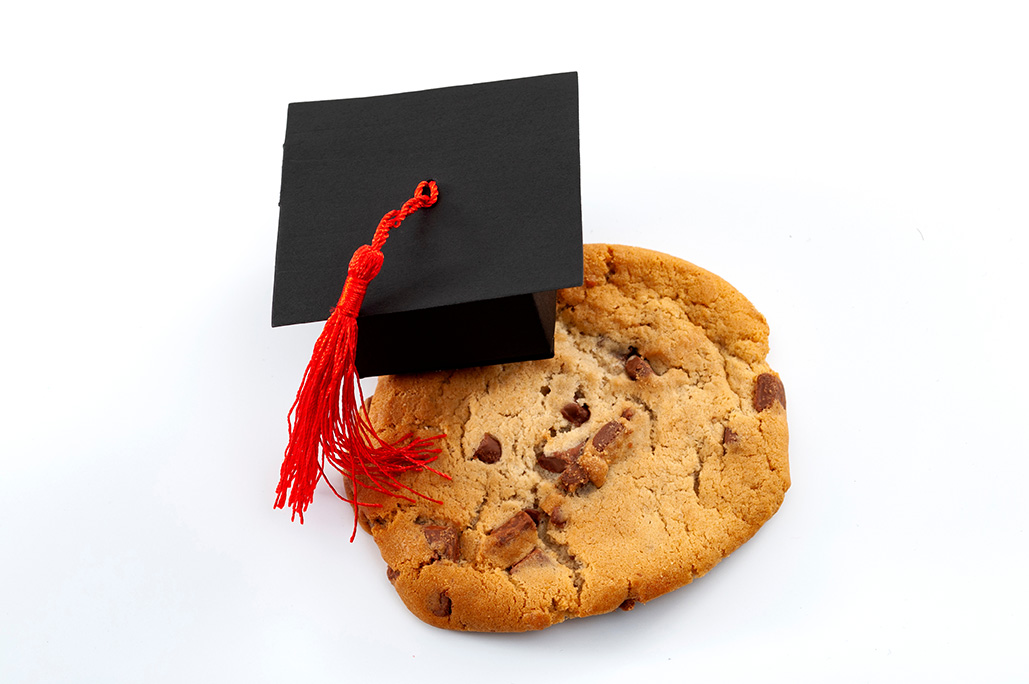 Chocolate chip cookie with a miniature mortarboard on it, isolated on white background.