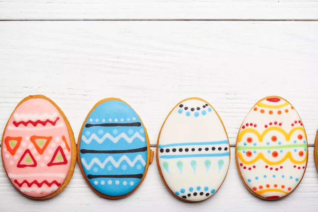 A row of four easter egg shaped sugar cookies, painted with edible watercolor patterns for Easter, against a white wooden background.