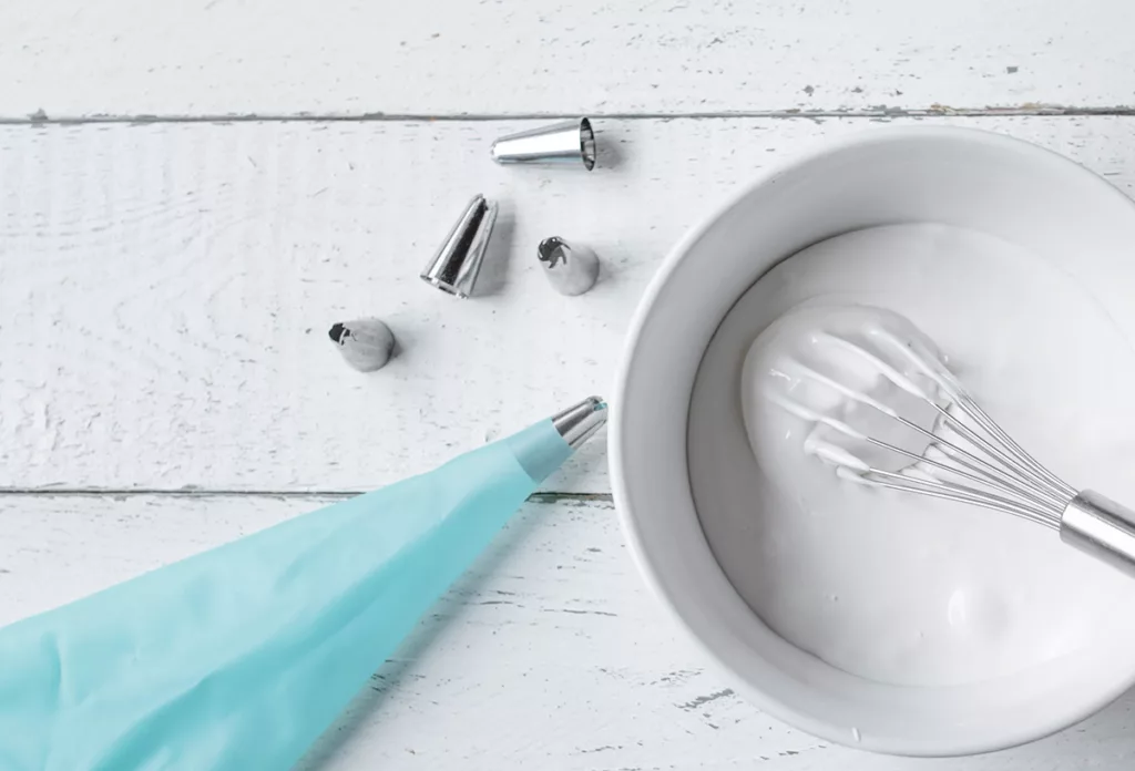 A bowl filled with white royal icing against a white wooden background, with a whisk and a blue frosting pipette.
