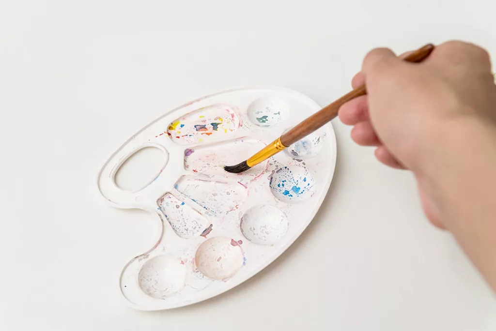 A hand dipping a food-safe paint brush into a palette of edible watercolor paints against a white background