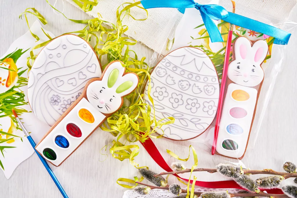 Two egg-shaped sugar cookies covered in white royal icing next to edible watercolors, ready to be decorated for Easter.